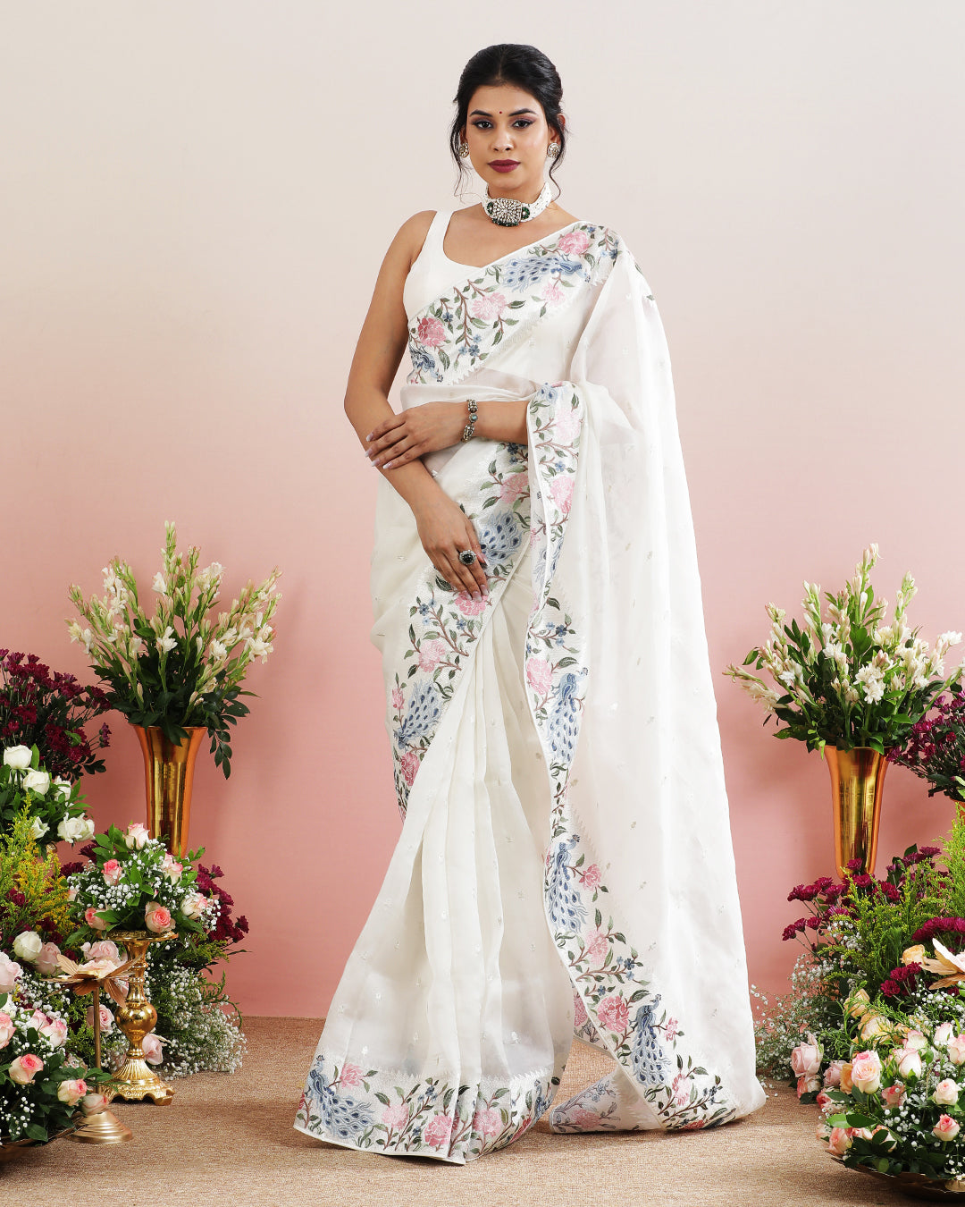 A woman modeling a white organza saree with multicolored floral embroidery along the border, standing against a backdrop of flowers.