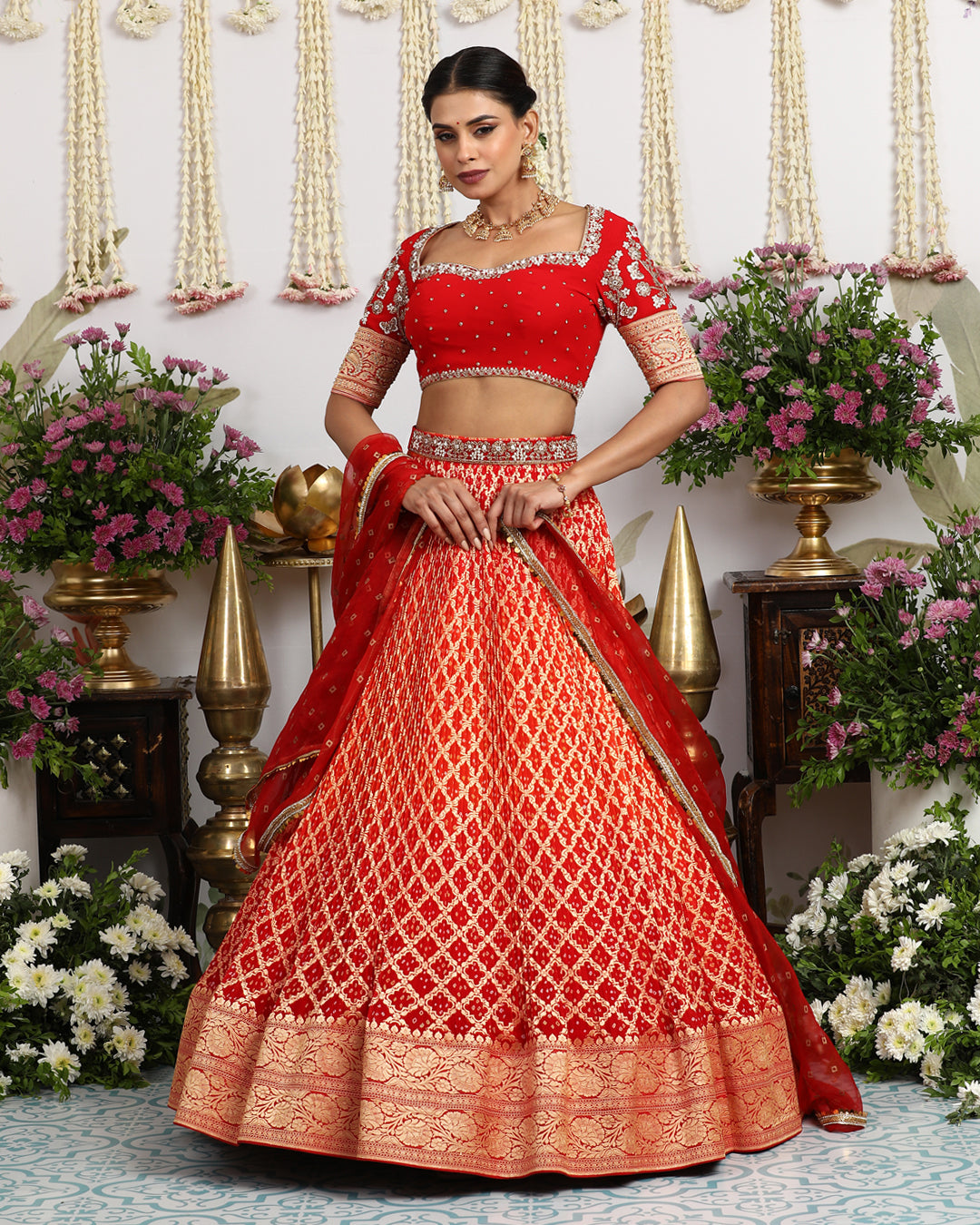 A model wearing a radiant red lehenga set with a geometric pattern, a sweetheart neckline blouse, and a matching red dupatta, set against a backdrop with floral decorations.